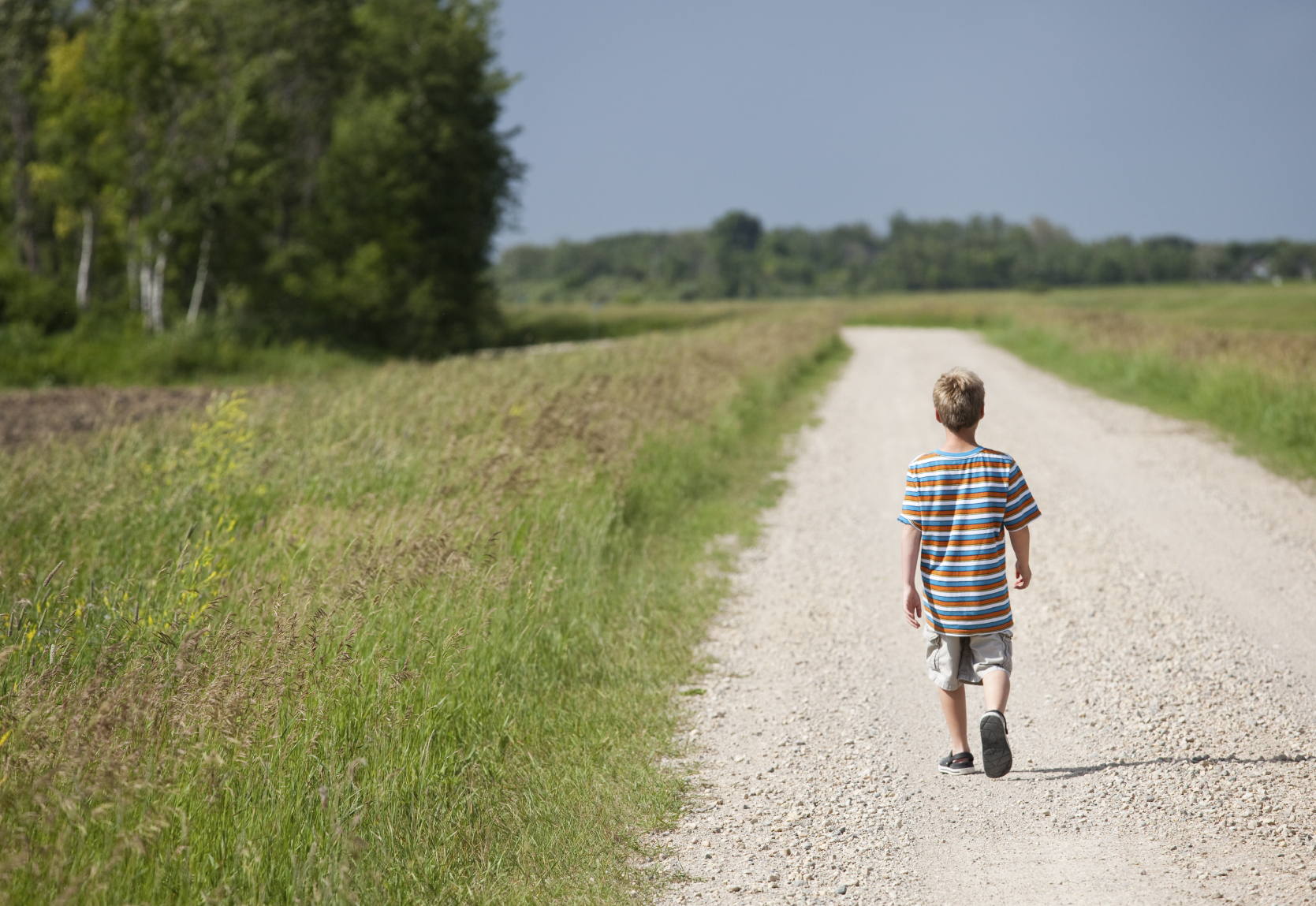 A young boy walks alone down a gravel road in the countryside.